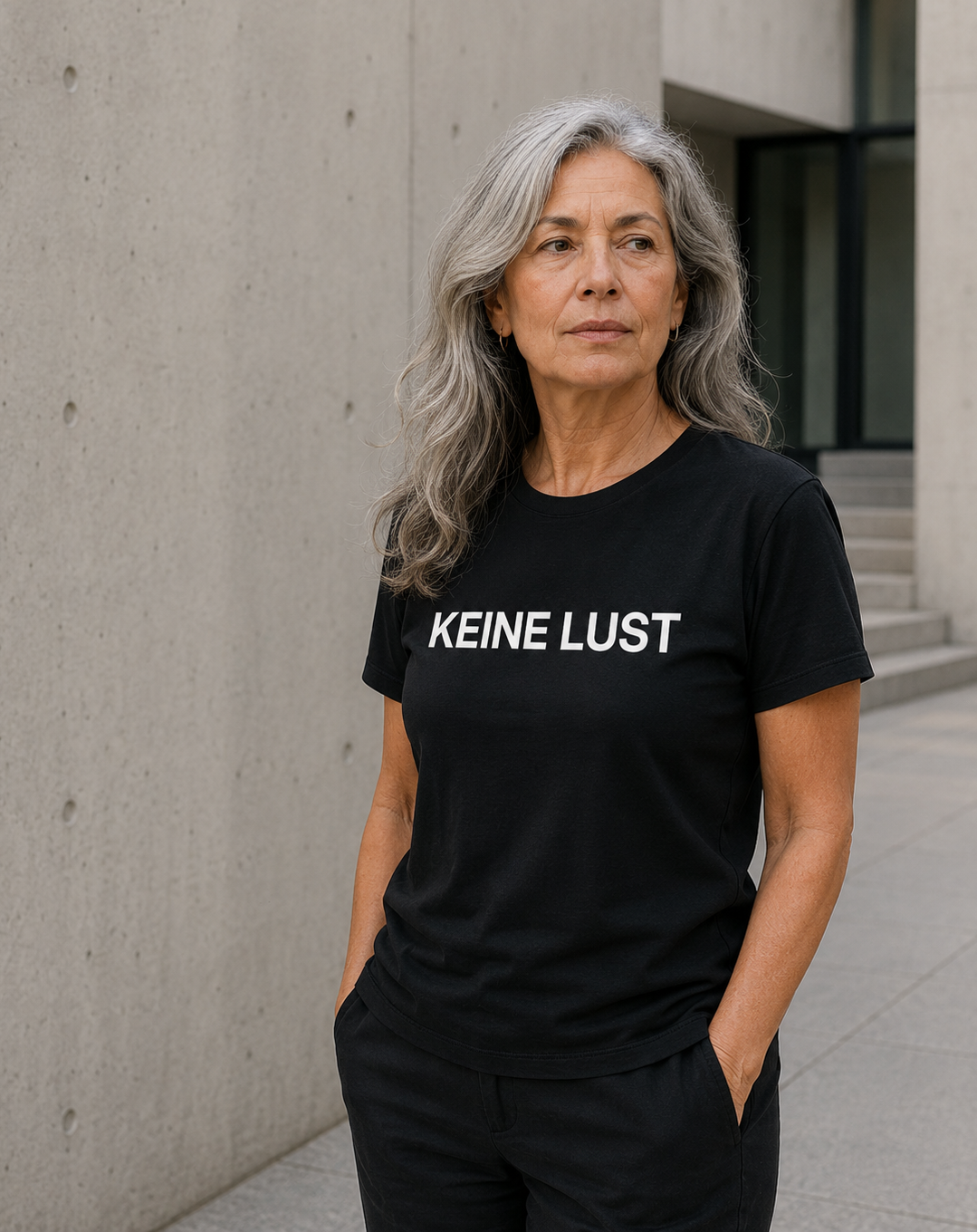 Mature woman wearing black KEINE LUST statement shirt in minimalist concrete setting.